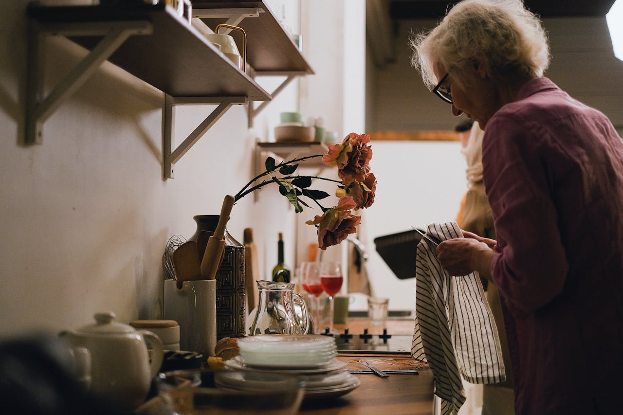 Elderly woman engaged in dish drying in a cozy, homey kitchen setting with shelves and flowers.
